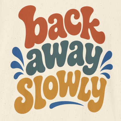 An off-white short-sleeve t-shirt with a retro-style graphic reading 'back away slowly' in orange, dark teal, and mustard yellow lettering, displayed on a clean white background.