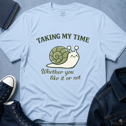 A light blue graphic t-shirt laid flat on a textured gray-blue surface, surrounded by navy sneakers, a denim jacket, and a notebook. The t-shirt features a cartoon snail with a green and cream shell and the text 'TAKING MY TIME' and 'Whether you like it or not' in olive green.