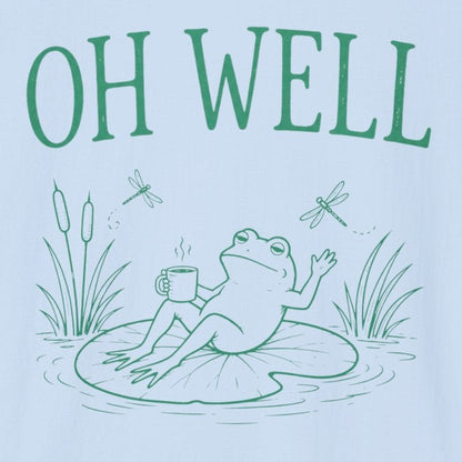 A light blue short-sleeve crewneck t-shirt featuring a green graphic print of a frog sitting on a lily pad with the text 'OH WELL' above it