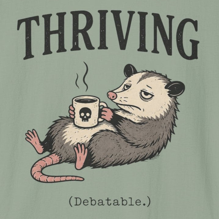 A sage green short-sleeve t-shirt with a graphic of an opossum holding a coffee mug under the word 'THRIVING' in black serif letters, on a plain white background.