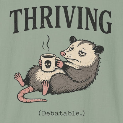 A sage green short-sleeve t-shirt with a graphic of an opossum holding a coffee mug under the word 'THRIVING' in black serif letters, on a plain white background.