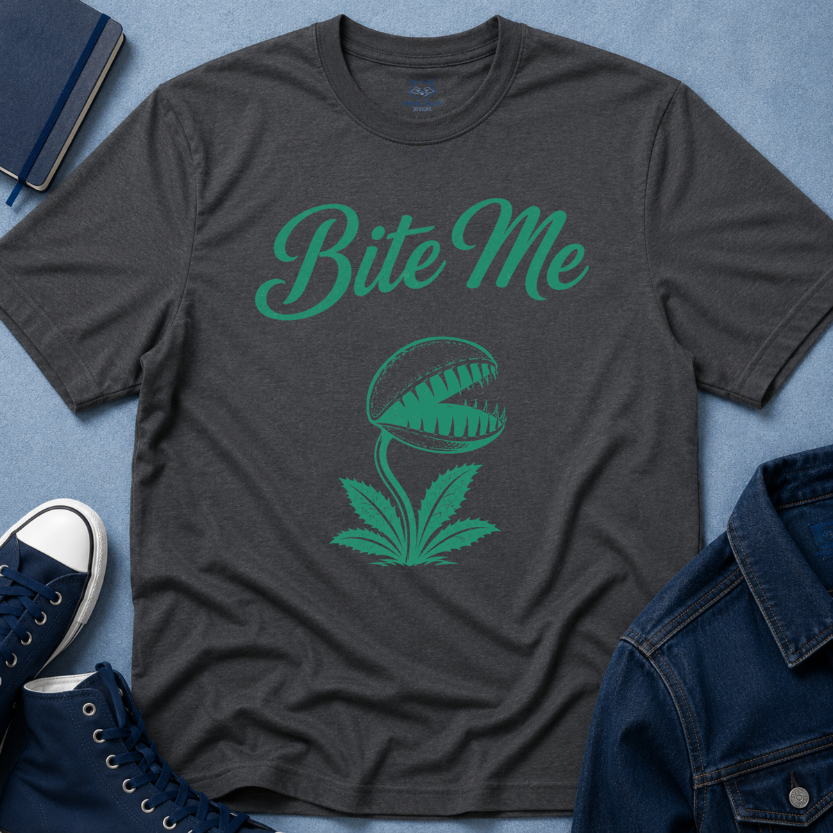 A dark charcoal gray t-shirt with a green graphic of a Venus flytrap and the text 'Bite Me' in green script. The shirt is styled in a flat-lay arrangement on a textured blue background, surrounded by navy blue sneakers, a denim jacket, and a notebook.