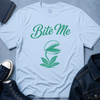 A light blue crewneck t-shirt with a green graphic of a Venus flytrap and the text 'Bite Me' in green script. The shirt is presented in a styled flat-lay arrangement with navy blue sneakers, a denim jacket, and a notebook on a textured gray background.