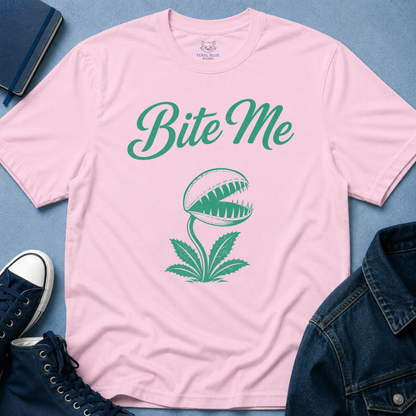 A light pink short-sleeve t-shirt with a green graphic of a Venus flytrap and the text 'Bite Me' in green script. The shirt is styled in a flat-lay arrangement on a blue textured background, surrounded by a navy blue denim jacket, a navy notebook, and navy sneakers.