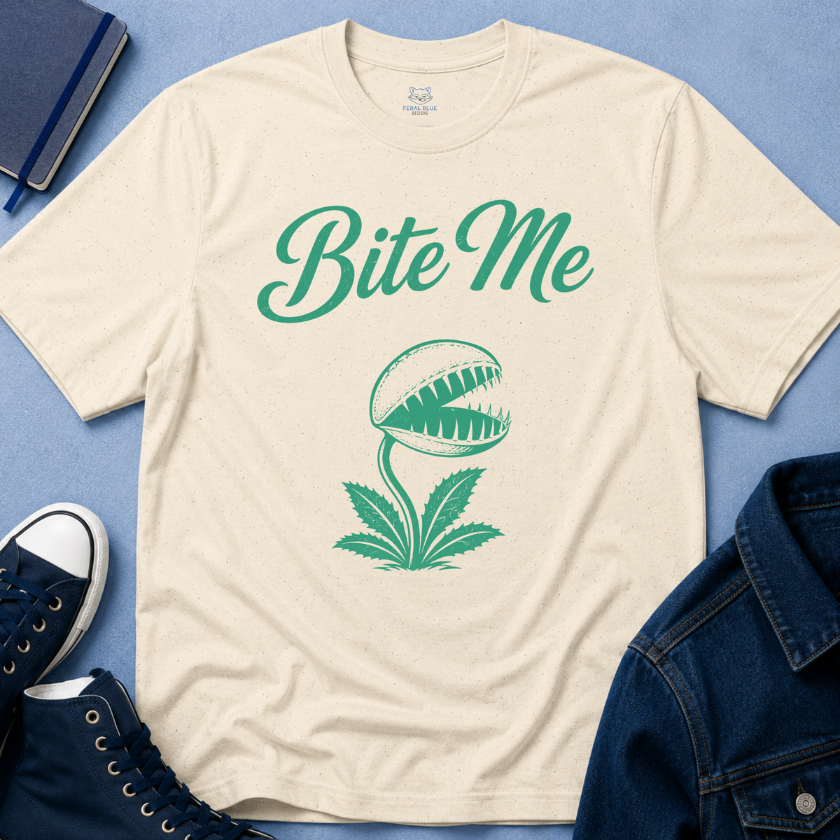 A cream-colored t-shirt with a green graphic of a Venus flytrap and the text 'Bite Me', styled in a flat-lay arrangement with a blue denim jacket, navy sneakers, a notebook, and a blue textured background.