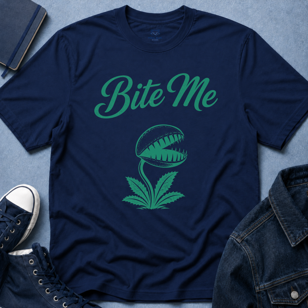 A navy blue crewneck t-shirt with a green graphic of a carnivorous plant and the text 'Bite Me', styled in a flat lay with a denim jacket, sneakers, and a notebook on a blue textured background.
