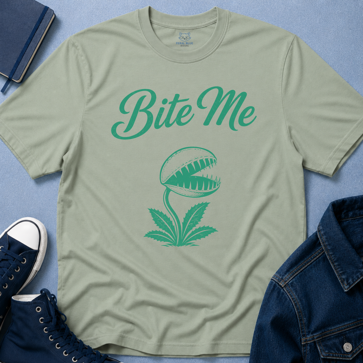 A sage green crewneck t-shirt with a green graphic of a carnivorous plant and the text 'Bite Me', styled in a flat-lay with a denim jacket, navy sneakers, and a notebook on a blue textured background.