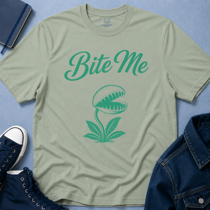 A sage green crewneck t-shirt with a green graphic of a carnivorous plant and the text 'Bite Me', styled in a flat-lay with a denim jacket, navy sneakers, and a notebook on a blue textured background.