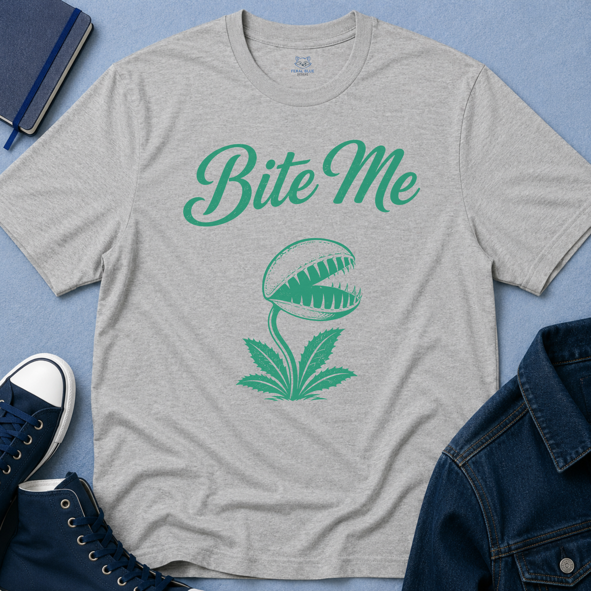 A heather gray short-sleeve t-shirt with a green graphic of the text 'Bite Me' above a Venus flytrap design, styled in a flat-lay with a blue denim jacket, navy sneakers, and a notebook on a blue background.