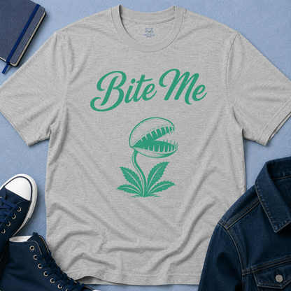 A heather gray short-sleeve t-shirt with a green graphic of the text 'Bite Me' above a Venus flytrap design, styled in a flat-lay with a blue denim jacket, navy sneakers, and a notebook on a blue background.